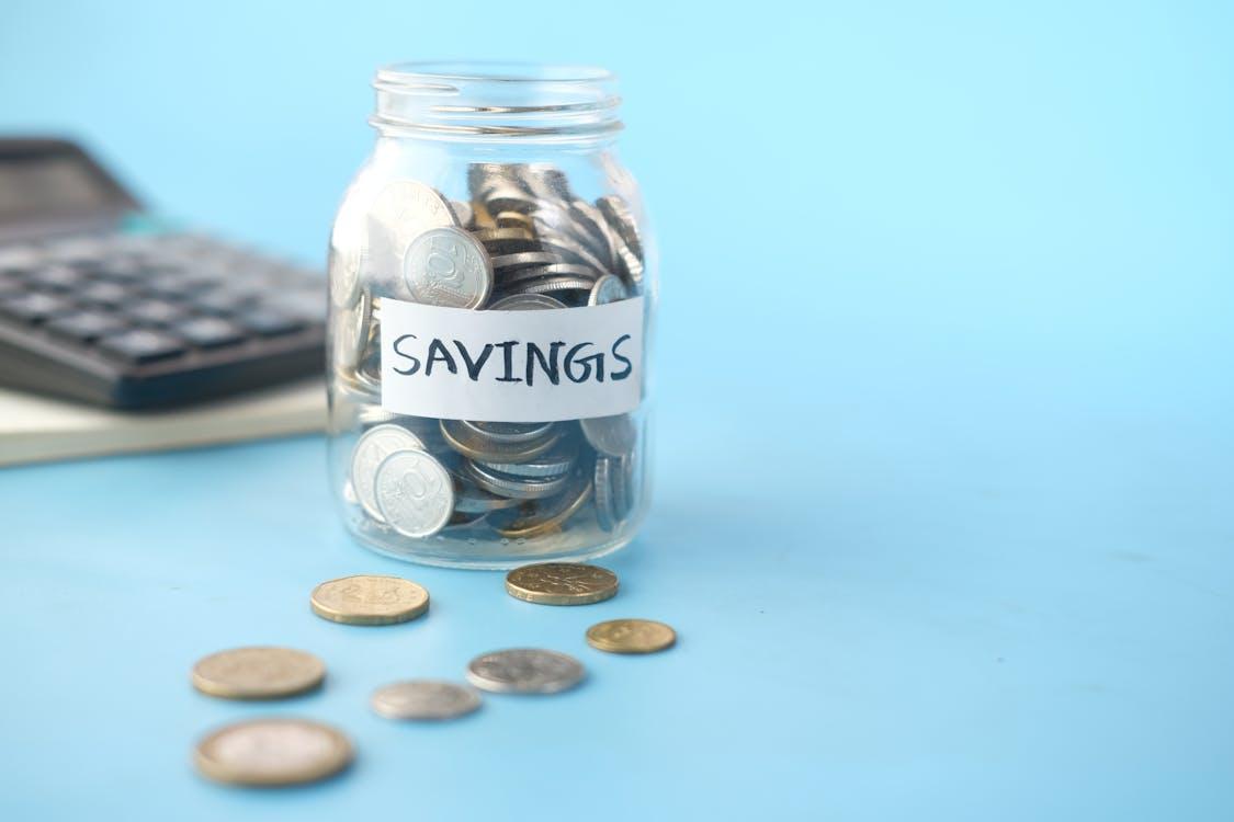 Free Glass jar labeled 'Savings' filled with coins, beside a calculator on a blue background. Stock Photo